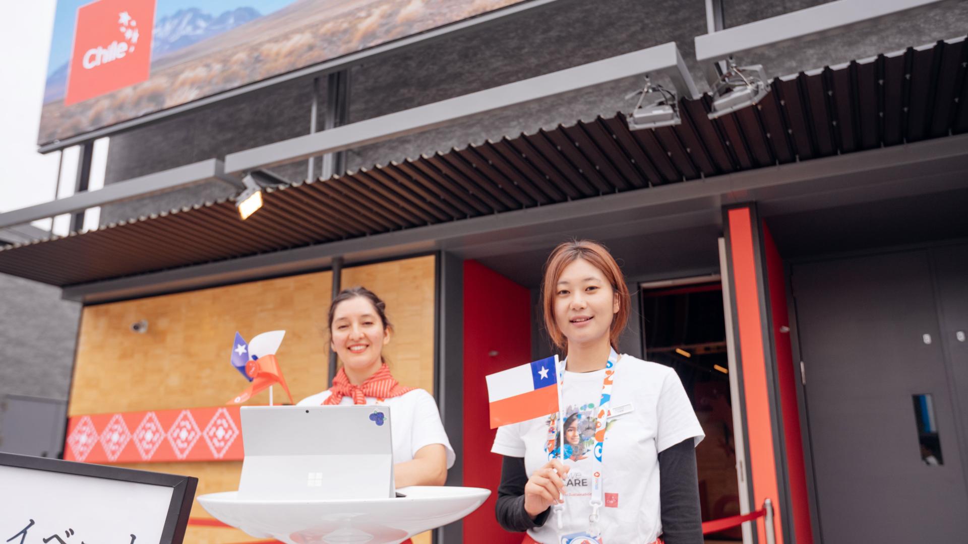 Dos mujeres con bandera chilena dan la bienvenida al pabellón de Chile en Osaka, Japón