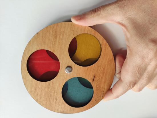 Mano sostiene circunferencia de madera con tres agujeros cubiertos por material transparente en colores rojo, verde y amarillo.
